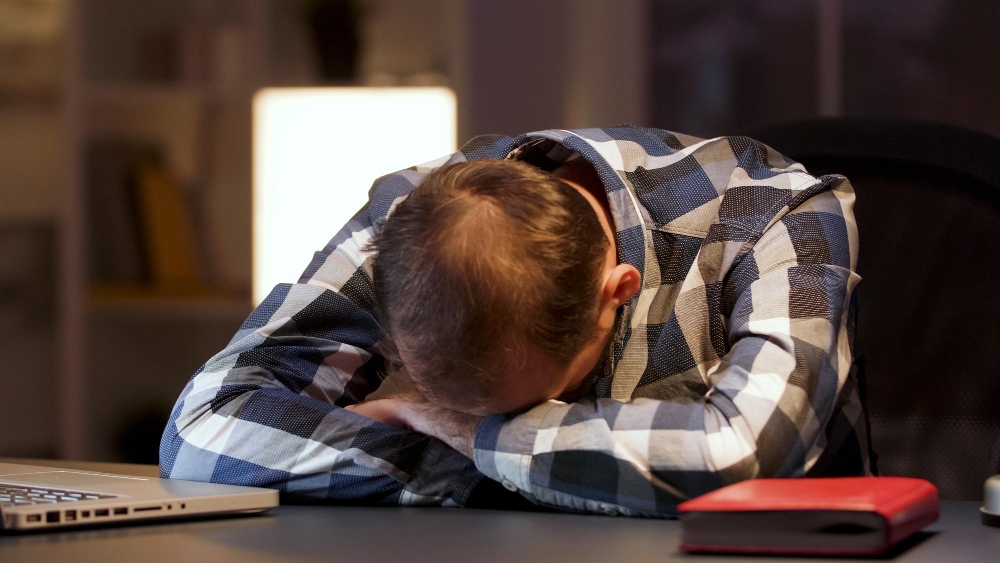 Bărbat epuizat cu capul pe birou lângă laptop